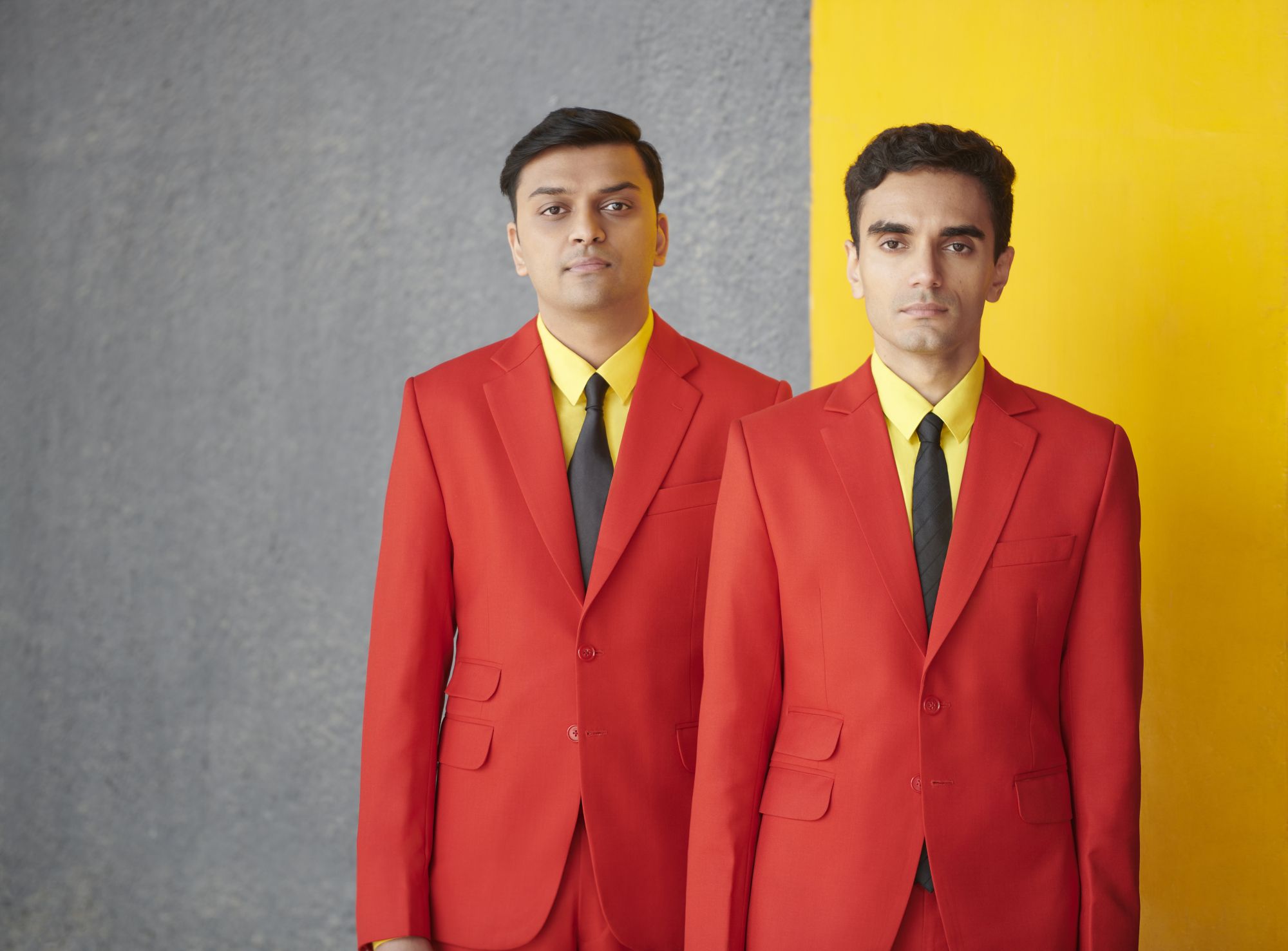 Photograph of Parekh & Singh in bright red suits. Parekh & Singh are wearing bright red suits, yellow shirts and black ties. The background is dark concrete and a painted yellow stripe.