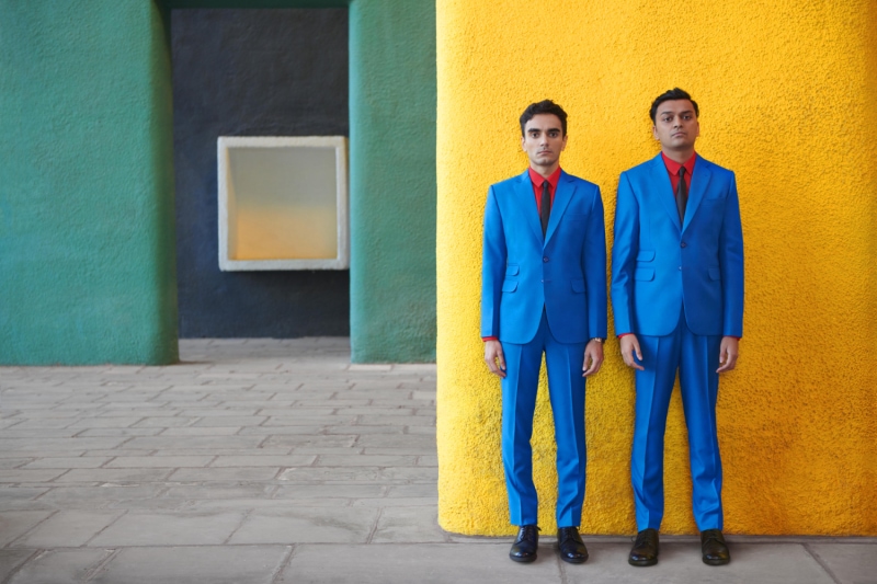 Photograph of Parekh & Singh in bright suits against a bright backdrop. Parekh & Singh wear bright blue suits with red shirts and black ties. The backdrop is the green and yellow columns of a building.