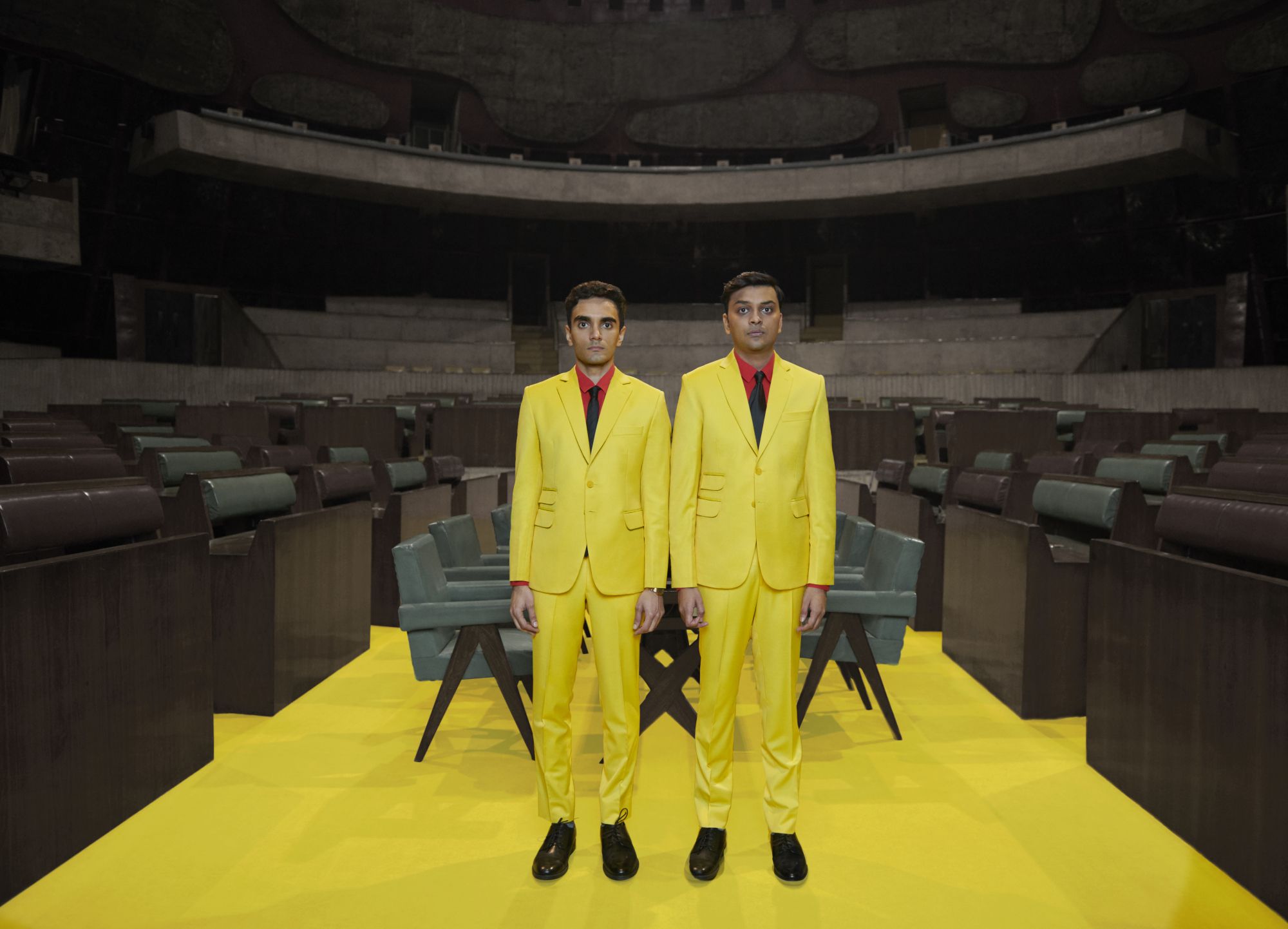 Photograph of Parekh & Singh wearing bright suits in an auditorium. Parekh & Singh are in bright yellow suits, red shirts, and black ties. They stand in an auditorium with a table and chairs behind them. The floor is bright yellow which matches their suits.