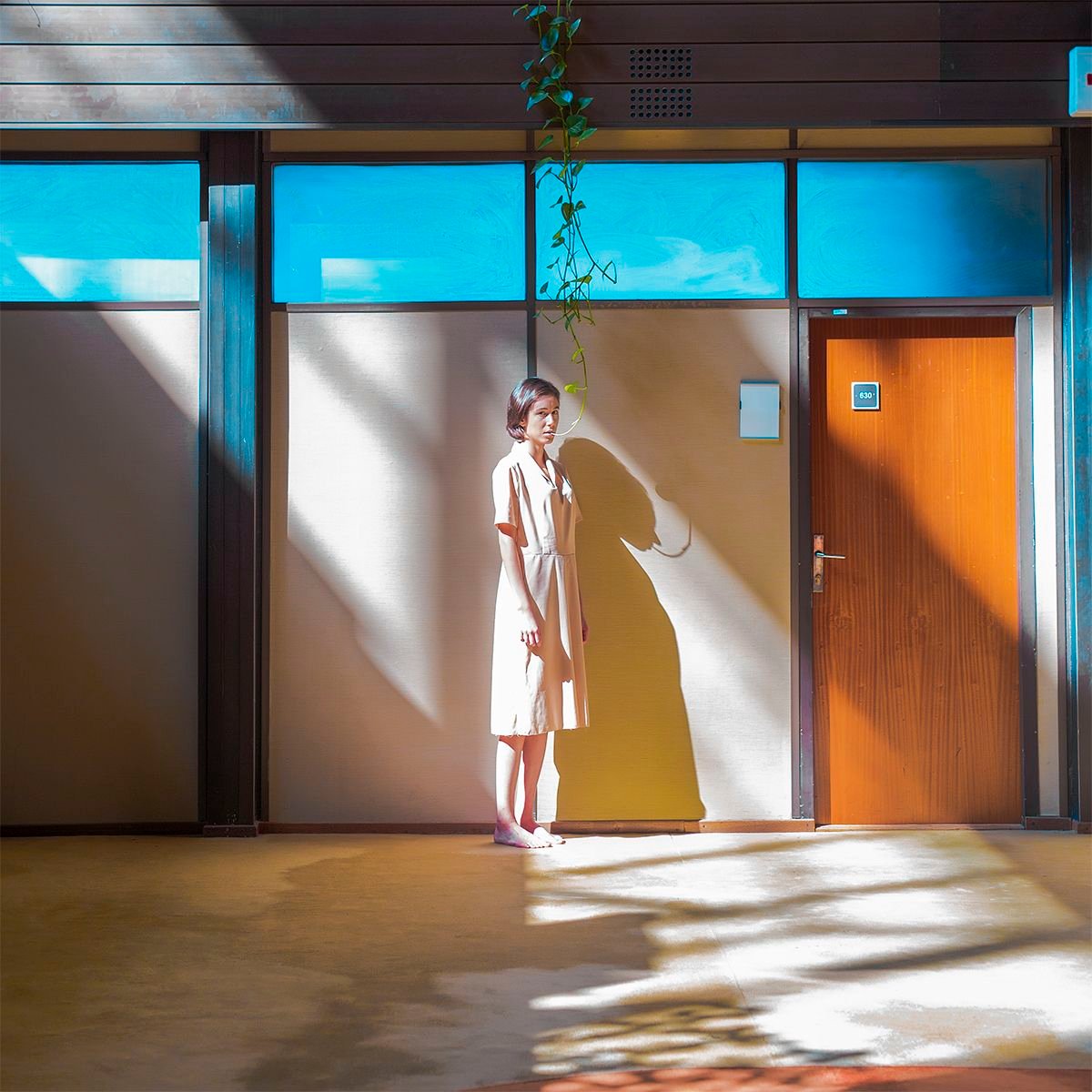 Photograph showing an empty office hallway where a woman stand still at the end. Her clothes blending into the beige walls, but above her hangs a plant, its tendrils connecting to her mouth