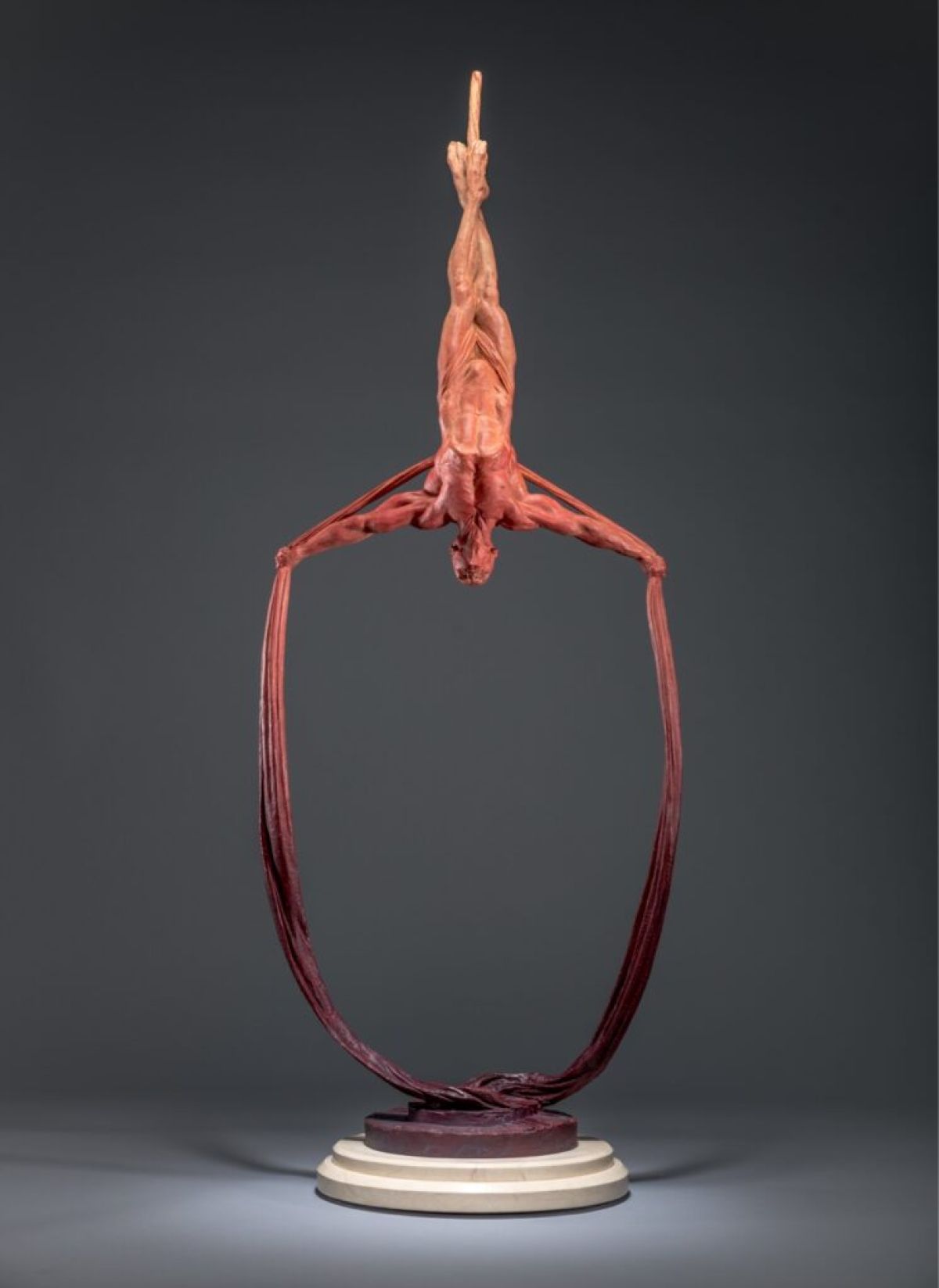Sculpture: An athletic man hangs upside down, his legs wrapped around silk, the material then extending down and around his outstretched arms and into a loop. It's from the bottom of this aerial silk loop that the sculpture rests. The sculpture is in a gradient linear red.