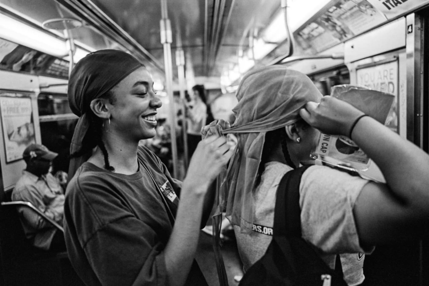 Black and white photograph showing two young women laughing and smiling on the train, one helps the other with their head scarf.