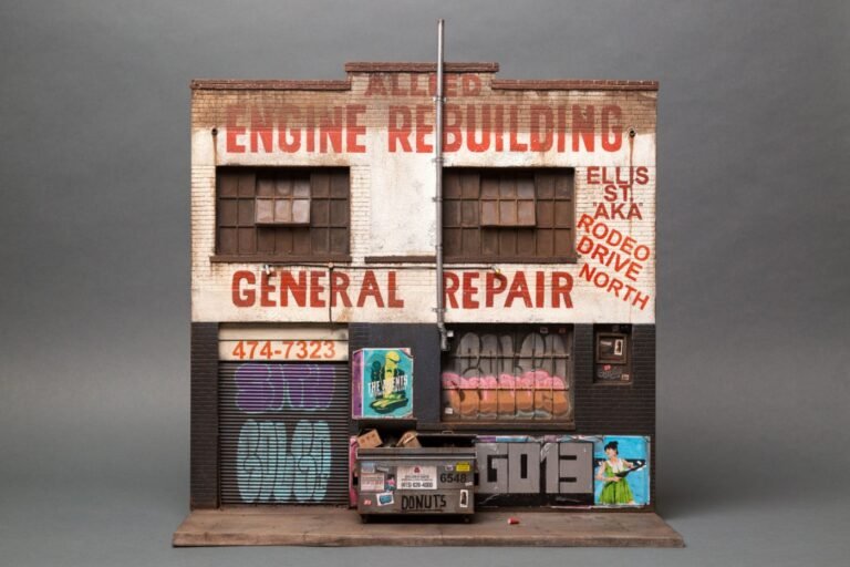 Miniature sculpture: A two storey building of a garage. Its shutters are down with graffiti tags across them. Meanwhile a dumpster stands in front with boxes peeping out from under the lid