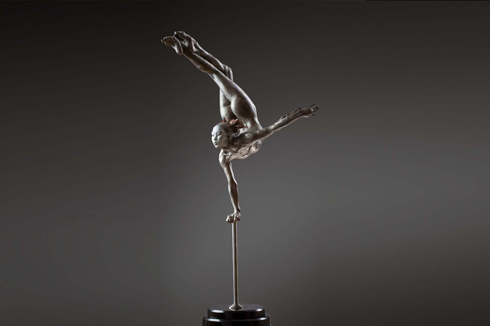 Sculpture: A narrow pole reaches up from the floor, its top covered by a small square rest. From this rest a girl hold herself upright by one arm, the other extended out to the side, while her legs are crossed and held up and behind her head - curving her form.