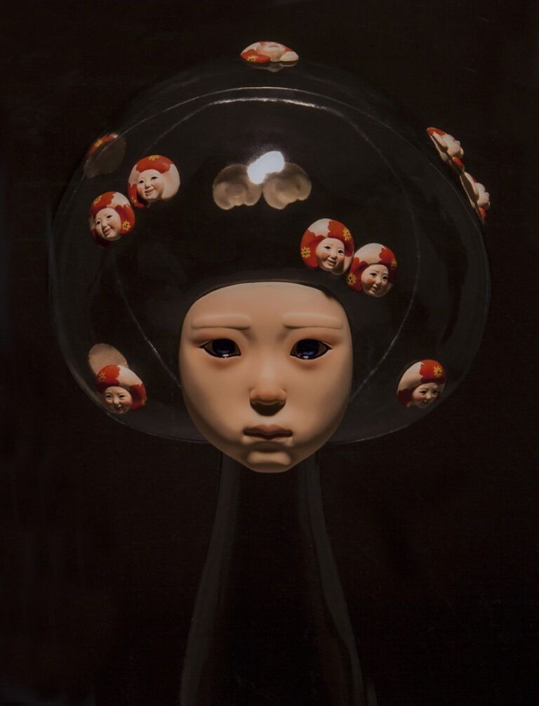 Transparent sculpture with solid face. The face is shown to be unhappy, while in their hair is multiple "selves" each showing a false emotion of happiness.