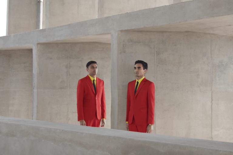 Photograph of Parekh & Singh in bright suits against open concrete sections. Parekh & Singh are in bright red suits, yellow shirts and black ties. The background is outdoors against a pale concrete open-section building.