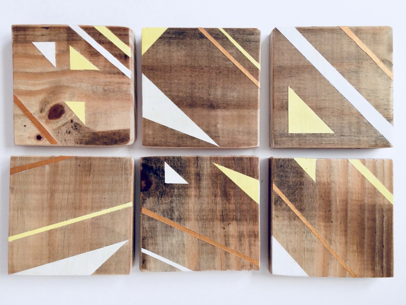 Six wooden coasters with painted geometric triangles and lines in white, yellow and gold.