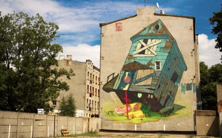 Photograph of a building with a large graffiti mural at one end. Its of a battered blue house propped up on a stick, and with a toxic slimed gammon below.