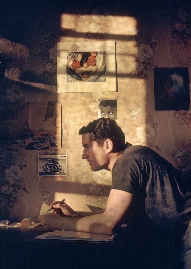 Photograph with a painterly appearance to it. The image is a self-portrait of the photographer Fred Herzog. The image shows a young Fred Herzog looking sideways and up from the table. Light has come in from the window which centres the image and creates a dramatic use of light to frame Herzog's face. It also shows artwork on the walls and a pen in Herzog's hand.