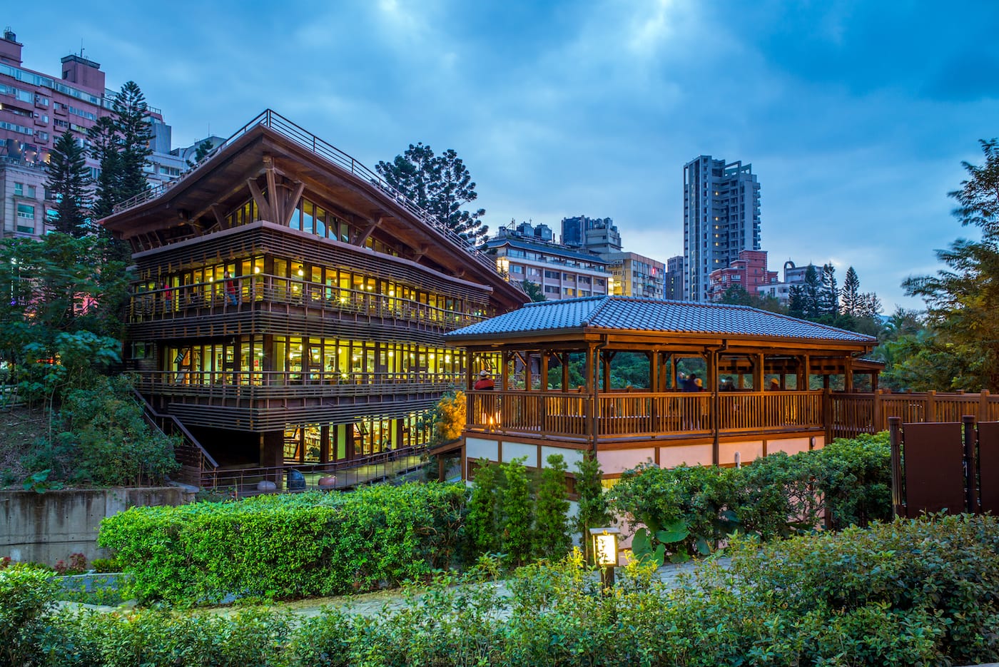 Two wooden buildings with lots of windows and wrap around terraces on each level. The buildings are set amongst greenery, while the background shows skyscrapers.